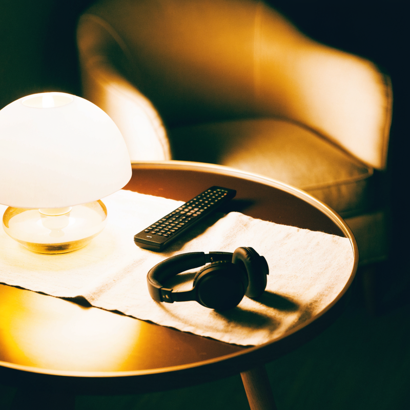 A television remote and headphones on a side table beside a warm lamp