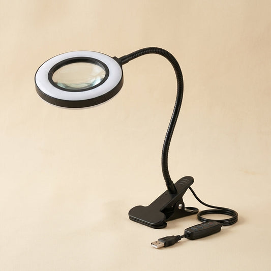 Clamp-on magnifying lamp with LED ring light, gooseneck arm and USB remote on a warm cream studio background