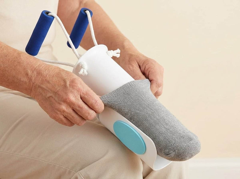 Two hands stretch a grey sock over the white cradle of a sock aid while seated.